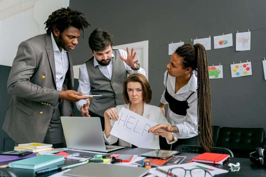Office scene with diverse colleagues around a stressed woman holding a sign that says 'HELP'. Focus on teamwork dynamics.