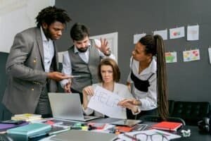 Office scene with diverse colleagues around a stressed woman holding a sign that says 'HELP'. Focus on teamwork dynamics.